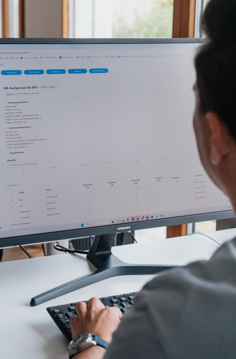 Eine Person sitzt vor einem Computerbildschirm und arbeitet im Publicjobs Login-Bereich. Ansicht der Bewerbungsdetailseite im ATS mit Bewerberprofil, Foto, persönlichen Angaben und Statusübersicht. Die Person ist von hinten zu erkennen.