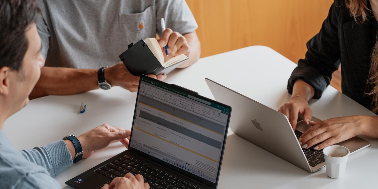 Mehrere Personen sitzen an einem Tisch und arbeiten gemeinsam mit Laptops und Notizen. Auf einem Bildschirm ist eine tabellarische Übersicht geöffnet, während eine weitere Person handschriftliche Notizen macht. Die Szene zeigt eine kollaborative Arbeitssituation im Büro.