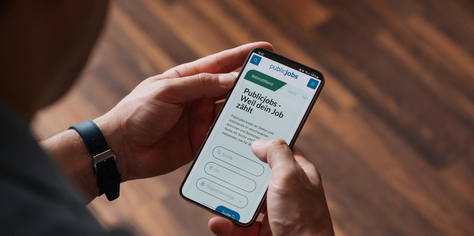 Person hält ein Smartphone mit geöffneter Website von publicjobs.ch, sichtbar ist die mobil optimierte Startseite der Stellenplattform mit Suchmaske und dem Claim «Weil dein Job zählt», passend zum Thema geräteübergreifende Darstellung von Stellenanzeigen.