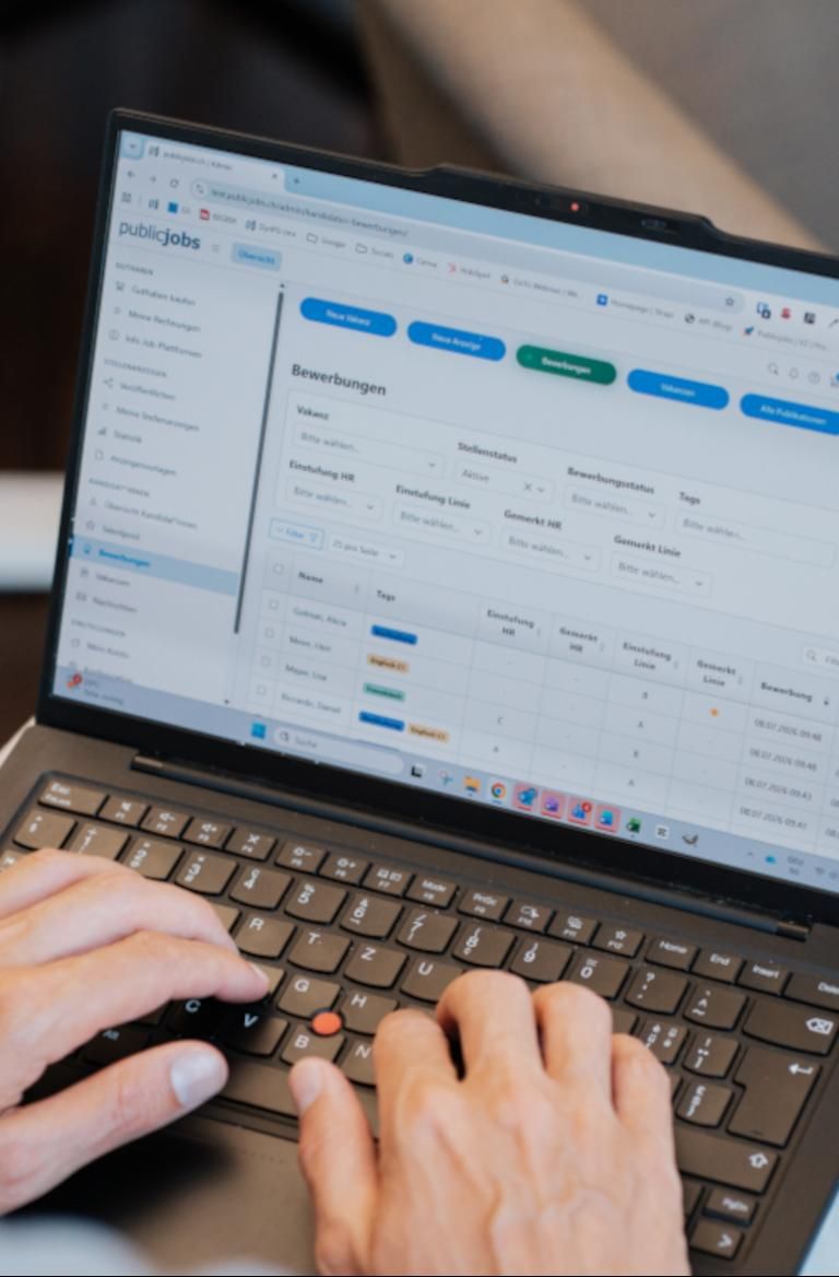 Person von hinten vor einem Computerbildschirm im Loginbereich von publicjobs.ch, sichtbar ist die Übersicht zur Verwaltung von Vakanzen als Teil der digitalen Rekrutierungslösung für sinnstiftende Organisationen.